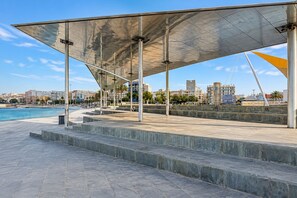 A waterfront promenade with a covered seating area, steps leading to a pool, and a cityscape in the background. - GuestReady - Sunset Canteras Sea Views (Las Palmas de Gran Canaria)