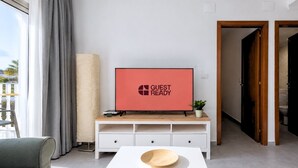 Una sala moderna con una televisión de pantalla plana mostrando 'GUEST READY', un mueble bajo blanco con cajones, un sofá gris y una mesa de centro de madera.