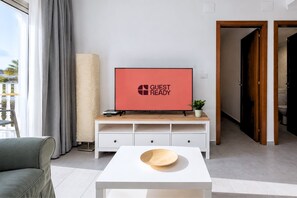 Ein modernes Wohnzimmer mit einem Flachbildfernseher, der 'GUEST READY' anzeigt, einem weißen Konsolentisch mit Schubladen, einem grauen Sofa und einem Holztisch.