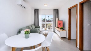 Una sala moderna con un sofá, una mesa de comedor con sillas, un televisor sobre un mueble de madera y un balcón con cortinas.