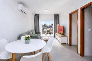 Ein modernes Wohnzimmer mit einer Couch, einem Esstisch mit Stühlen, einem Fernseher auf einem Holzboden und einem Balkon mit Vorhängen.
