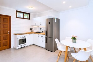 Une cuisine compacte avec des meubles blancs, un réfrigérateur en acier inoxydable, un plan de travail en bois et un petit coin repas avec des chaises et une table blanches.