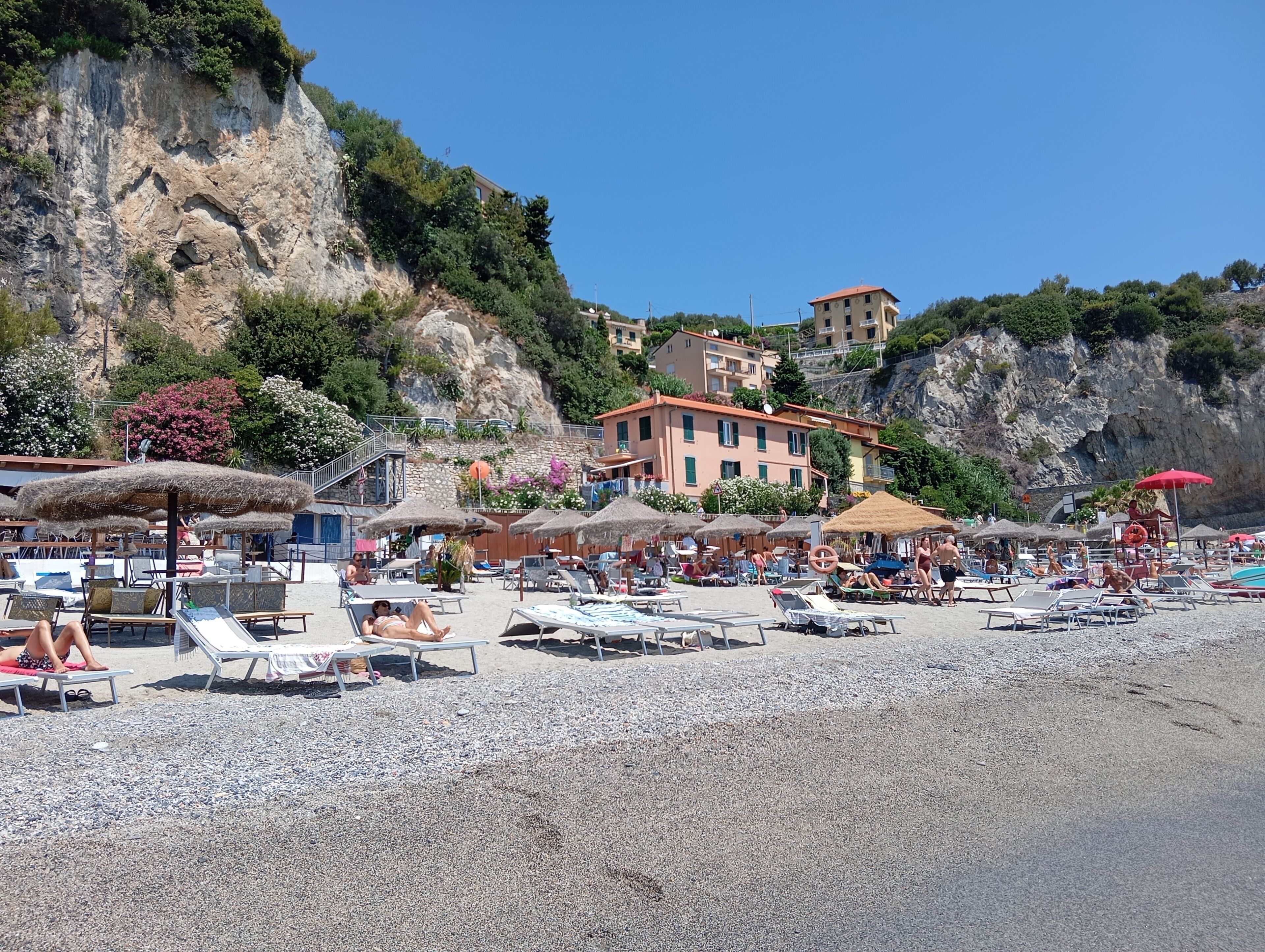 Une plage avec des chaises longues et des parasols, un bâtiment avec un toit rouge et, au loin, une falaise rocheuse.