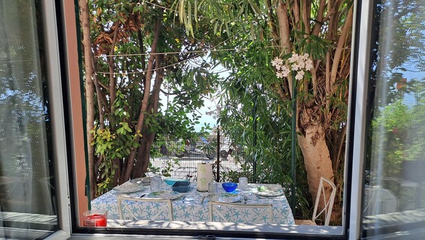 A window view of a dining area with a table set for two, surrounded by greenery and a tree.