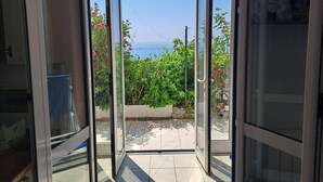 A view through a glass door leading to a patio with a tiled floor, a garden with green foliage, and a clear blue sky.