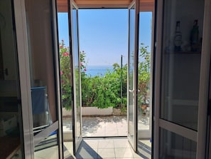 A view through a glass door leading to a patio with a tiled floor, a garden with green foliage, and a clear blue sky. - Orizzonte Blu Giardino sul Mare, Finale Ligure, on the beachfront (Finale Ligure)