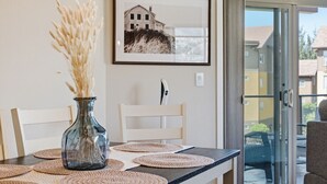 A dining table with placemats, a vase with dried plants, a framed picture on the wall, and a sliding glass door leading to an outdoor area. - 3BR Condo | Lake & Pool Views | Beach Resort (West Kelowna)