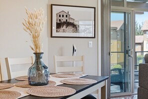 A dining table with placemats, a vase with dried plants, a framed picture on the wall, and a sliding glass door leading to an outdoor area. - 3BR Condo | Lake & Pool Views | Beach Resort (West Kelowna)