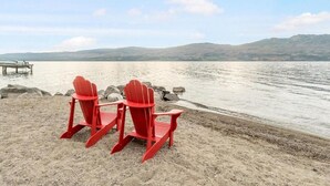 Two red Adirondack chairs on a sandy beach with a lake and mountains in the background. - 3BR Condo | Lake & Pool Views | Beach Resort (West Kelowna)