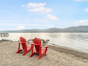 Two red Adirondack chairs on a sandy beach with a lake and mountains in the background. - 3BR Condo | Lake & Pool Views | Beach Resort (West Kelowna)