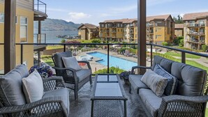 A balcony with wicker furniture, a pool, and a view of a mountain and other buildings. - 3BR Condo | Lake & Pool Views | Beach Resort (West Kelowna)