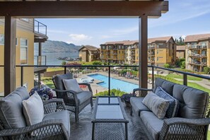 A balcony with wicker furniture, a pool, and a view of a mountain and other buildings. - 3BR Condo | Lake & Pool Views | Beach Resort (West Kelowna)