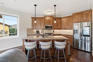 A kitchen with wooden cabinets, a stainless steel refrigerator, a granite countertop, and three bar stools. - 3BR Condo | Lake & Pool Views | Beach Resort (West Kelowna)