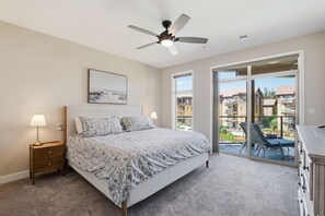 A bedroom with a bed, bedside lamps, a ceiling fan, a large window, and a balcony with a view of other buildings. - 3BR Condo | Lake & Pool Views | Beach Resort (West Kelowna)