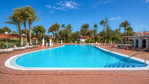 A clear blue swimming pool surrounded by lounge chairs and palm trees, with a tiled deck and a clear sky. - GuestReady - Sun Club Hammock Relax (San Bartolomé de Tirajana)
