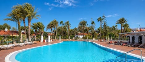 A clear blue swimming pool surrounded by lounge chairs and palm trees, with a tiled deck and a clear sky.