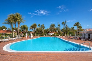 A clear blue swimming pool surrounded by lounge chairs and palm trees, with a tiled deck and a clear sky. - GuestReady - Sun Club Hammock Relax (San Bartolomé de Tirajana)