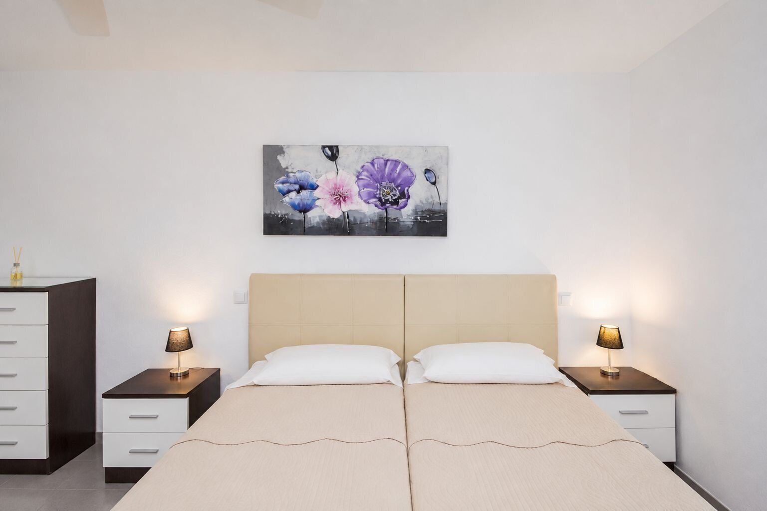 A bedroom with a double bed, two bedside tables with lamps, a nightstand, a dresser, and a wall-mounted painting.