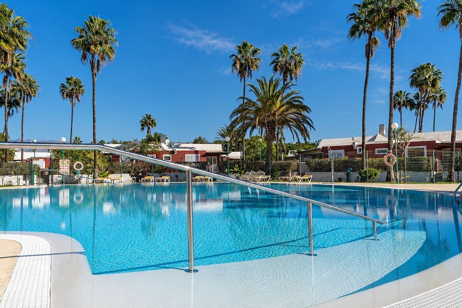 A clear blue pool with a metal safety railing, surrounded by palm trees and buildings.