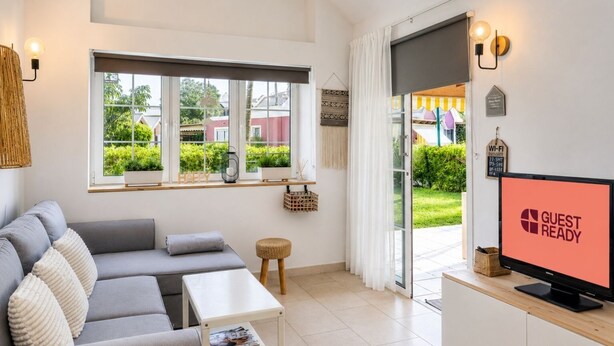 A modern living room with a grey sofa, a small table, a TV displaying 'GUEST READY', and a large window with a view of greenery and buildings. - GuestReady - Los Tunos Family Pool (Maspalomas)