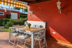 Eine Terrasse mit einem Holztisch und weißen Stühlen, eine rote Wand mit der Nummer 76 sowie ein kleiner Topfpflanze.
