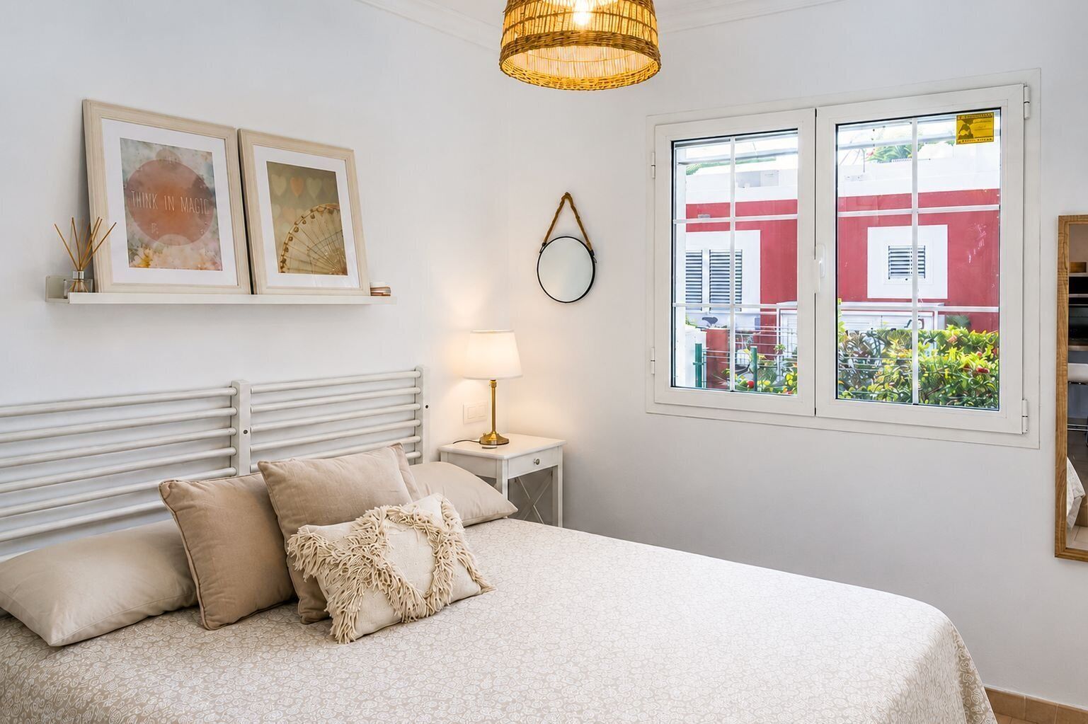 Ein Schlafzimmer mit einem Bett, einer Nachttischlampe, einem Fenster mit Blick auf ein rotes Gebäude und zwei gerahmten Kunstwerken an der Wand.