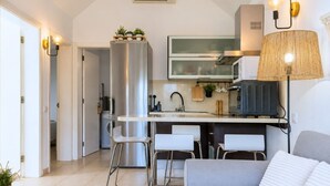 A modern kitchen with a stainless steel refrigerator, a microwave, and a built-in oven. There are two white bar stools and a grey sofa with a white cushion. - GuestReady - Los Tunos Family Pool (Maspalomas)
