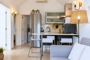 Eine moderne Küche mit einem Edelstahlkühlschrank, einer Mikrowelle und einem eingebauten Backofen. Zwei weiße Barhocker und ein graues Sofa mit einem weißen Kissen.