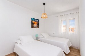 A room with two beds, a painting, a hanging light fixture, and a window with blinds.