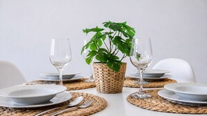 Mesa posta com pratos, talheres e dois copos de vinho, com um jogo americano de fibra natural e um vaso com planta ao centro.