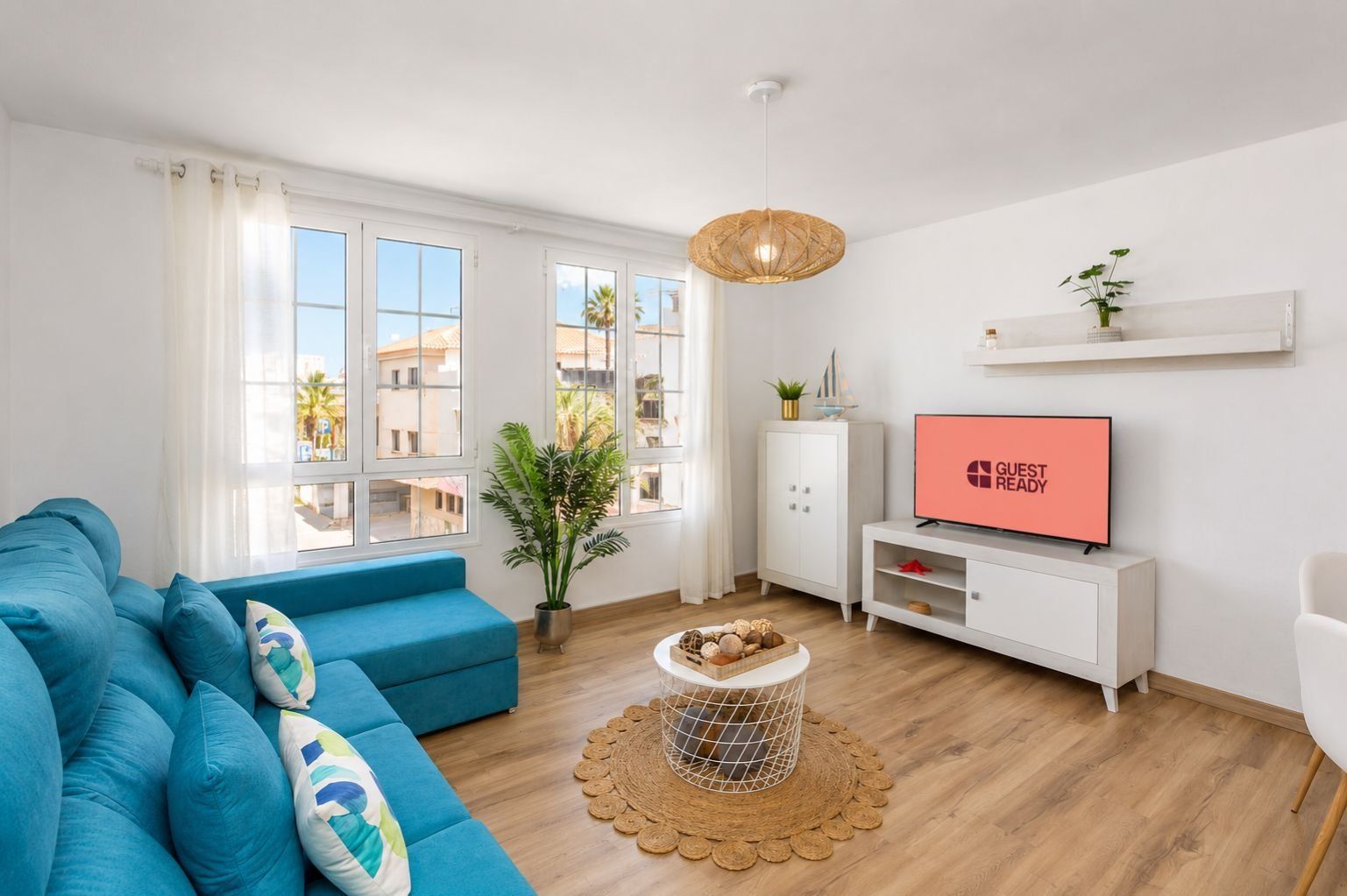 A modern living room with a blue sofa, a television displaying 'GUEST READY', a white cabinet, and a large window with a view of buildings and palm trees.