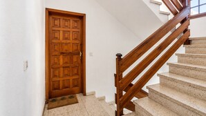 Uma porta de madeira com superfície texturizada, uma escada de madeira com corrimão e um tapete de entrada no chão.