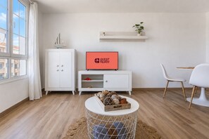 A modern living room with a white cabinet, a television displaying 'GUEST READY', a round coffee table with decorative items, and a view of the outside through a window.