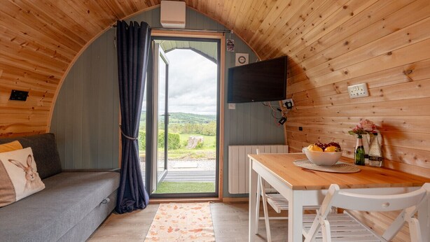 A cozy cabin interior with a sofa, dining table, and a view through a large door. - Greendale Glamping - Sunrise Pod (No Hot Tub) (Chatburn)