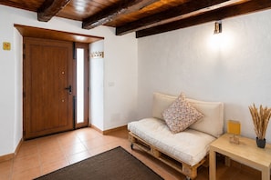 Ein Zimmer mit Holztür, einem Sofa mit einem Blumenkissen, einem kleinen Tisch mit einer Lampe und einer Vase mit getrockneten Pflanzen.