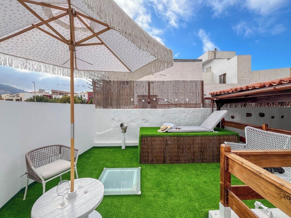 A rooftop patio with artificial grass, a table with a glass top, a wicker chair, a wooden bench, and a large umbrella.