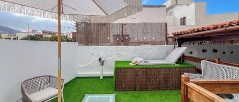 A rooftop patio with artificial grass, a table with a glass top, a wicker chair, a wooden bench, and a large umbrella.