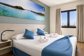 A hotel room with a bed, a nightstand, a desk lamp, a painting of a beach, and a window with curtains. - GuestReady - Luxury Villa with Pool (Antigua)