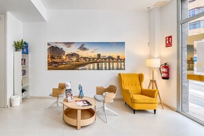 Um saguão de hotel moderno com uma pintura da paisagem urbana, uma poltrona amarela, uma mesa redonda de madeira e uma estante de livros.