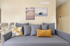 Un canapé gris agrémenté de plusieurs coussins, une chaise blanche et deux œuvres d’art fixées au mur, représentant un paysage désertique et un bateau.