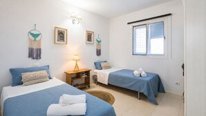 Ein Zimmer mit zwei Betten, einem Nachttisch mit Lampe, Wandleuchten, gerahmten Bildern und einem Fenster mit Jalousien.