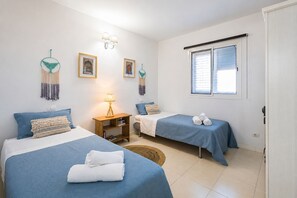 A room with two beds, a bedside table with a lamp, wall-mounted lights, framed pictures, and a window with blinds. - GuestReady - La Palmita Agaete with Pool (Agaete)
