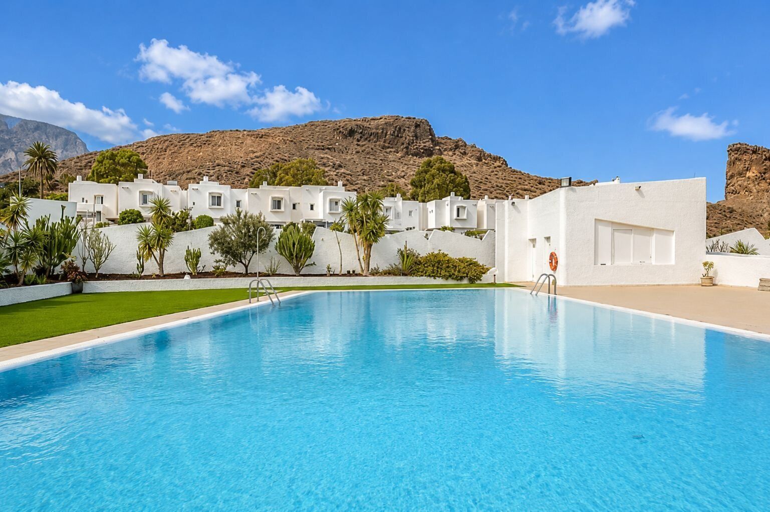 A swimming pool with a clear blue water surface, surrounded by white buildings with multiple windows, set against a mountainous backdrop.