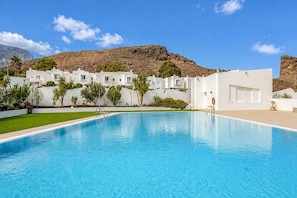 A swimming pool with a clear blue water surface, surrounded by white buildings with multiple windows, set against a mountainous backdrop. - GuestReady - La Palmita Agaete with Pool (Agaete)