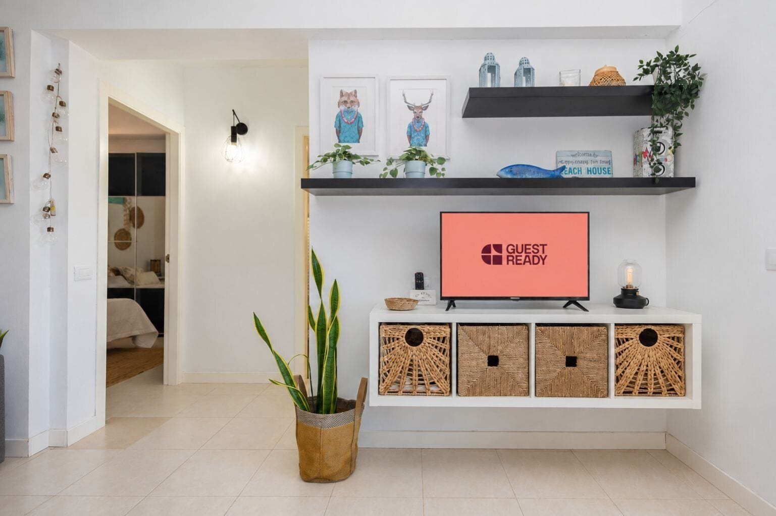 Ein modernes Wohnzimmer mit einem Flachbildfernseher, der 'GUEST READY' anzeigt, einem an der Wand montierten Regal mit Dekoration und einer Topfpflanze.