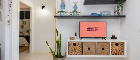 A modern living room with a flat-screen TV displaying 'GUEST READY,' a wall-mounted shelf with decorative items, and a potted plant.