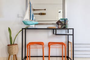 A room with a black metal console table, two orange wire mesh chairs, a small potted plant, a decorative sailboat, and a mirror reflecting a door and a wooden object. - GuestReady - La Palmita Agaete with Pool (Agaete)