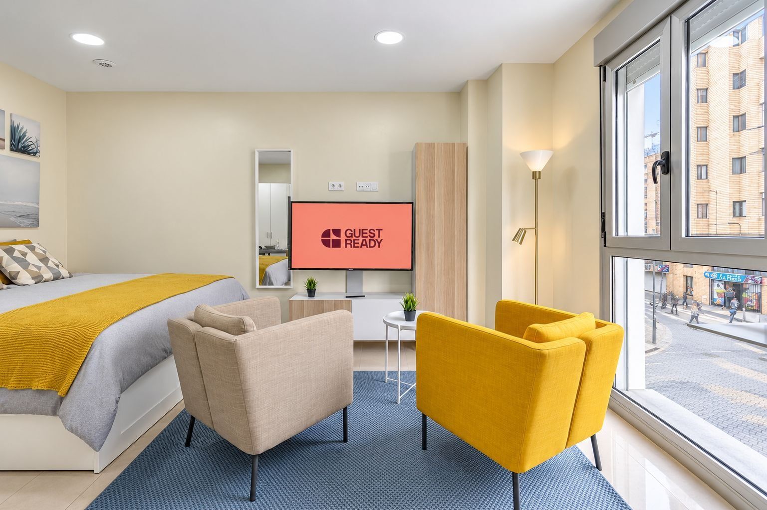 Ein Hotelzimmer mit einem Bett, einem Fernseher der 'GUEST READY' anzeigt, einem Schreibtisch, einem Stuhl und einem großen Fenster mit Blick auf die Straße.