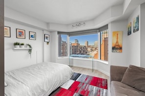 Ein Schlafzimmer mit großem Fenster, einem Bett, einer Couch und Blick auf die Stadt.