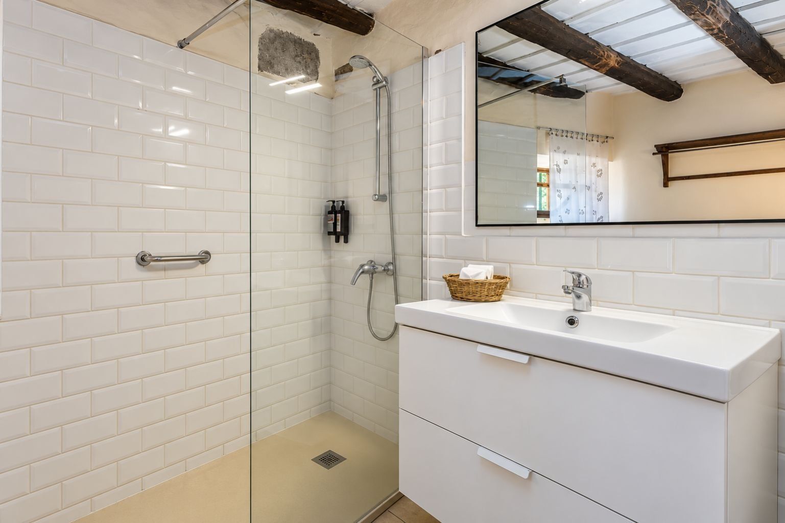 Un baño con ducha envuelta en cristal, paredes de azulejos blancos, un lavabo blanco con una cesta y un travesaño de madera.